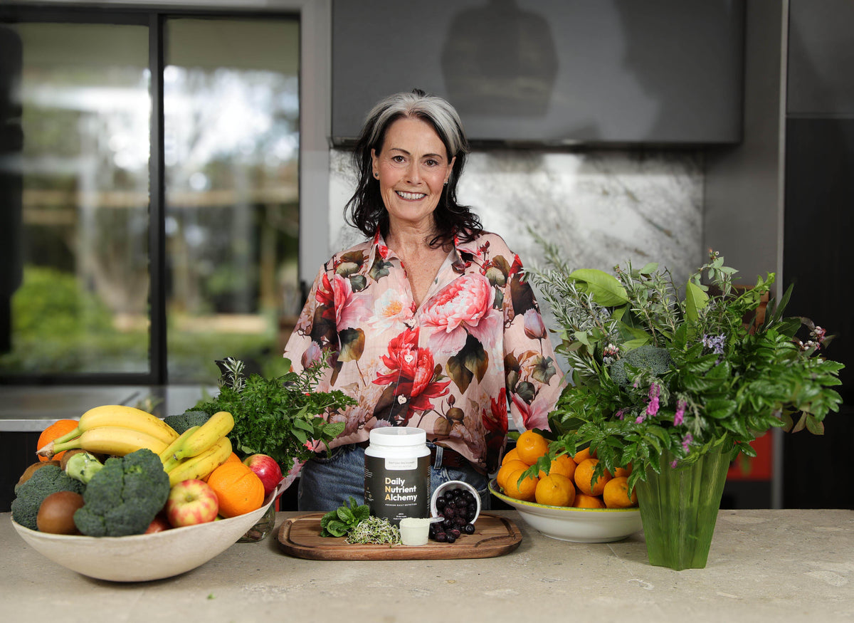 A smiling woman with gray hair stands behind a kitchen counter displaying a jar of 'Plant Based Protein Powder' surrounded by fresh fruits, vegetables, and flowers in a modern kitchen setting.