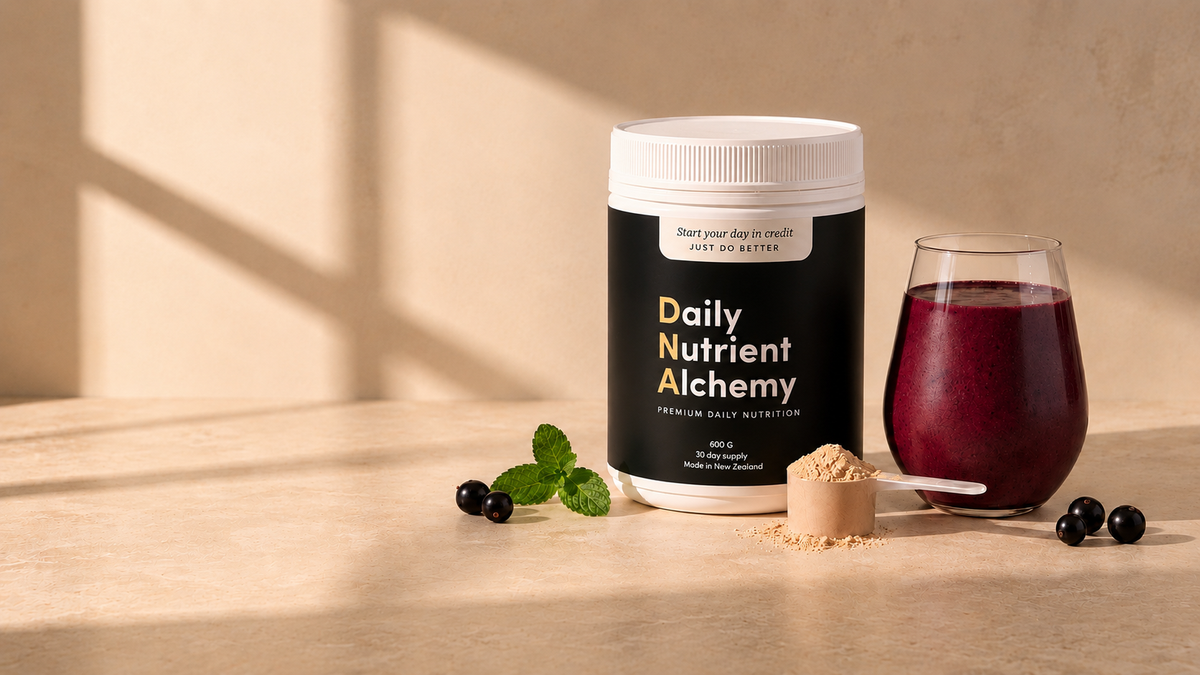 A white tub of Daily Nutrient Alchemy powder with a black label stands on a beige surface beside a glass of dark berry drink, a scoop of powder, mint leaves, and blueberries in a warm, sunlit setting.