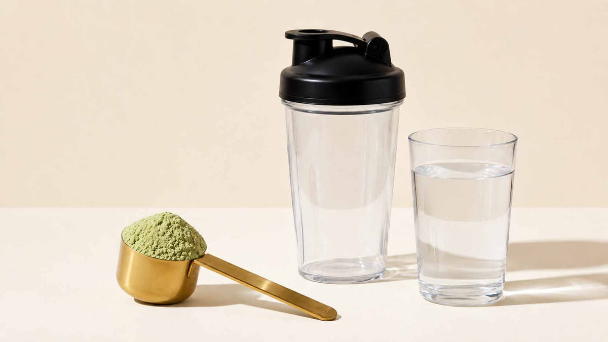 A clear shaker bottle with a black lid stands next to a glass of water and a gold scoop filled with green powder on a light beige background.